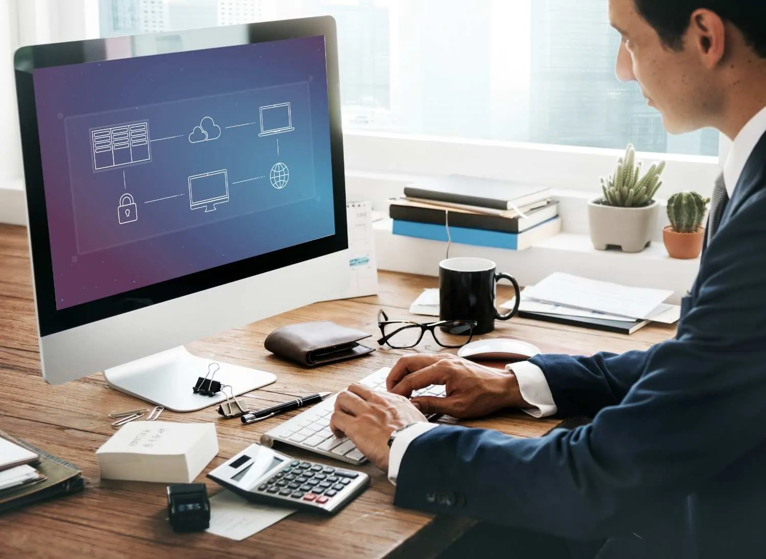 Man in a suit typing at a desktop computer showing cloud network icons, calculator on the desk