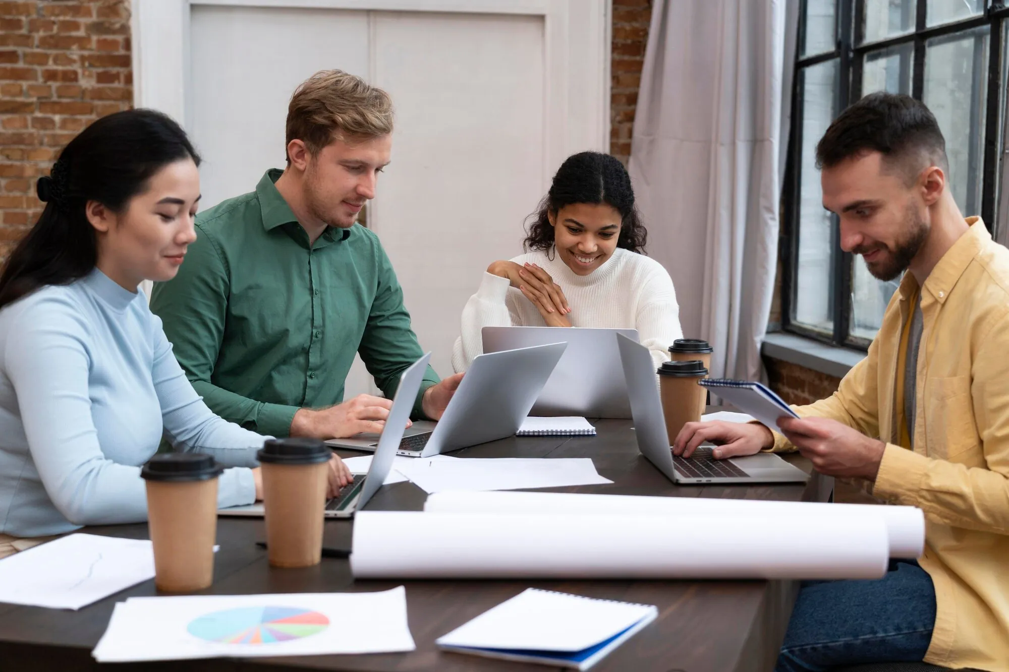 Stock photo of four colleagues working on laptops at a table with coffee cups and paper charts