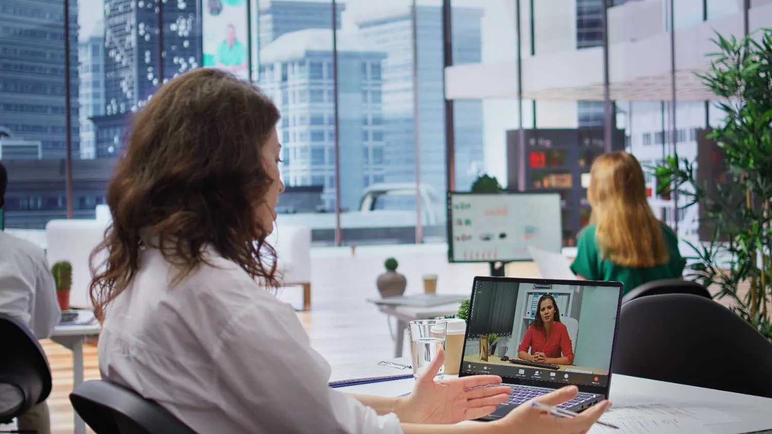 Woman on a video call, laptop screen showing another woman, in a bright city view office